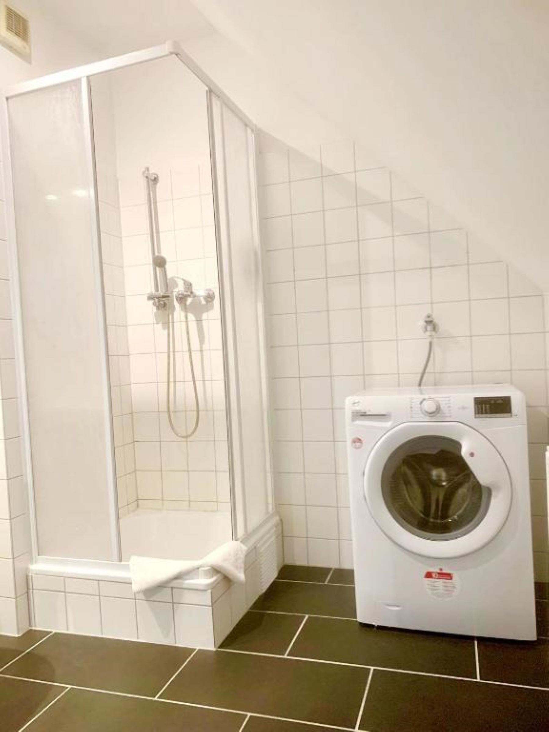 A corner bathroom with a glass-enclosed shower, a built-in washing machine, and dark brown tiled flooring.
