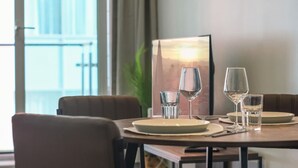 A dining table with four chairs, a framed picture on the wall, and a window with a view of a cityscape. - O2 & ExCeL! Modern 1 Bed Flat + Sofa Bed • Ideal for Conferences & Families (London)