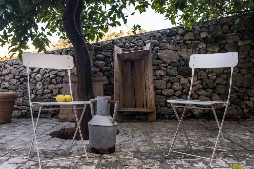 Twee witte klapstoelen met metalen frames, een houten deur met een landelijke uitstraling, en een stenen muur op de achtergrond.