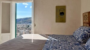 Une chambre à coucher avec un lit, une porte donnant sur un balcon, un tableau encadré au mur et une vue sur le paysage urbain.