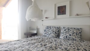 A bedroom with a bed, a bedside table, a lamp, a window with curtains, and a wall-mounted shelf with decorative items. - Casa do Forno - Rural Retreat in the Serra de São Mamede with Panoramic Views (Santo António das Areias)