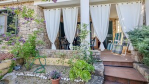 Una casa de pedra amb un porxo cobert, cortines blanques, mobles d'exterior i plantes en testos.