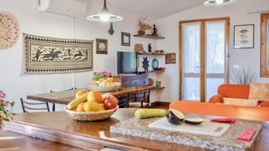 Una cuina amb una taula de menjador, un plat de fruita, una taixa de tallar i un televisor.