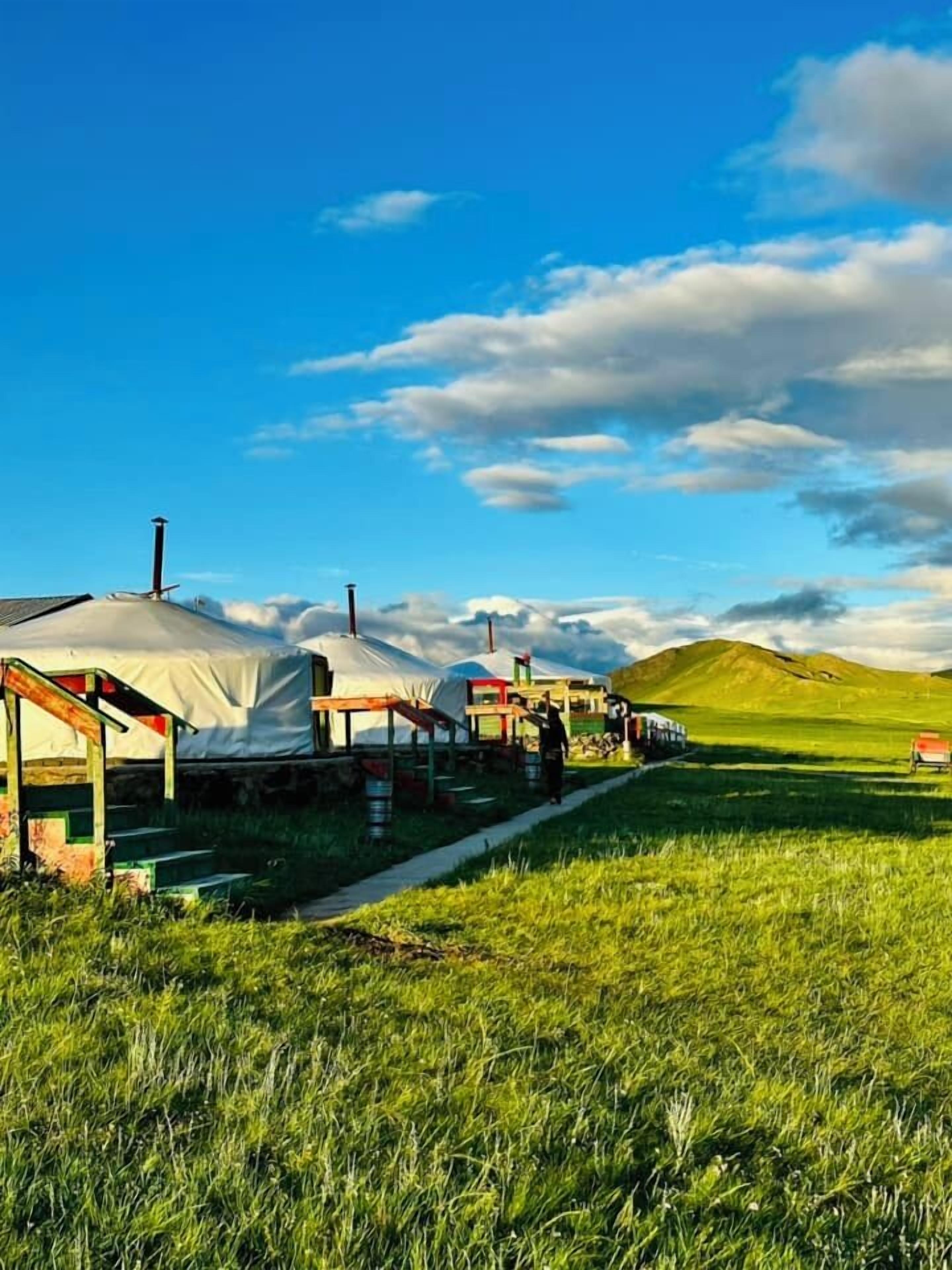 Yurts with a pathway leading to a grassy field and a hill in the background.