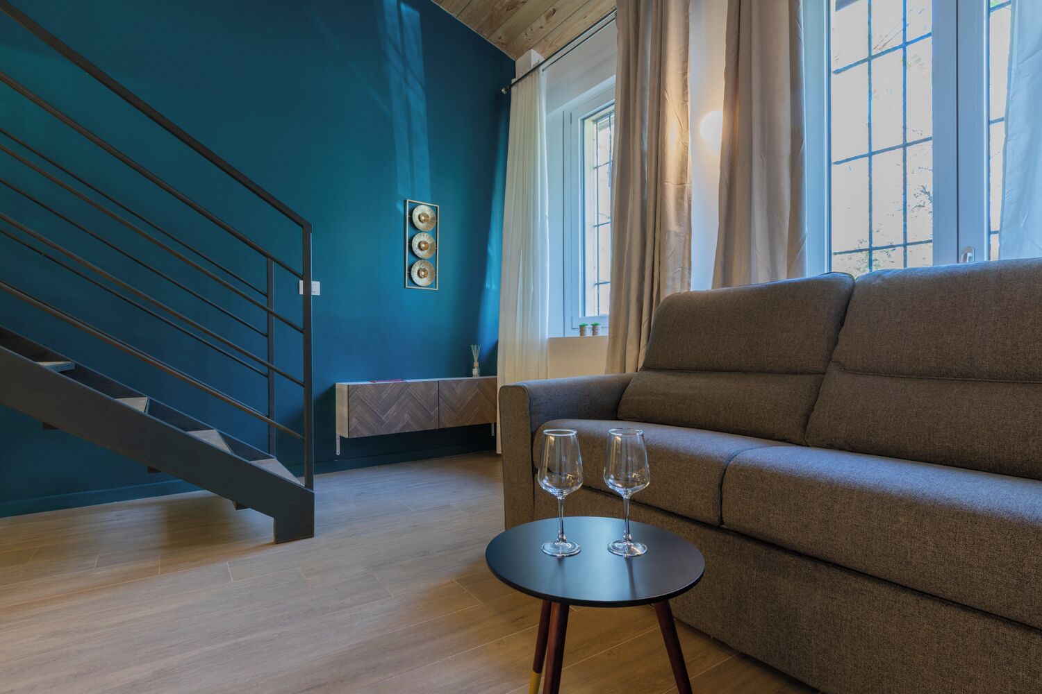 Un salon moderne avec un canapé, une petite table sur laquelle reposent deux verres à vin, un meuble au motif chevrons et un escalier adossé à un mur bleu.