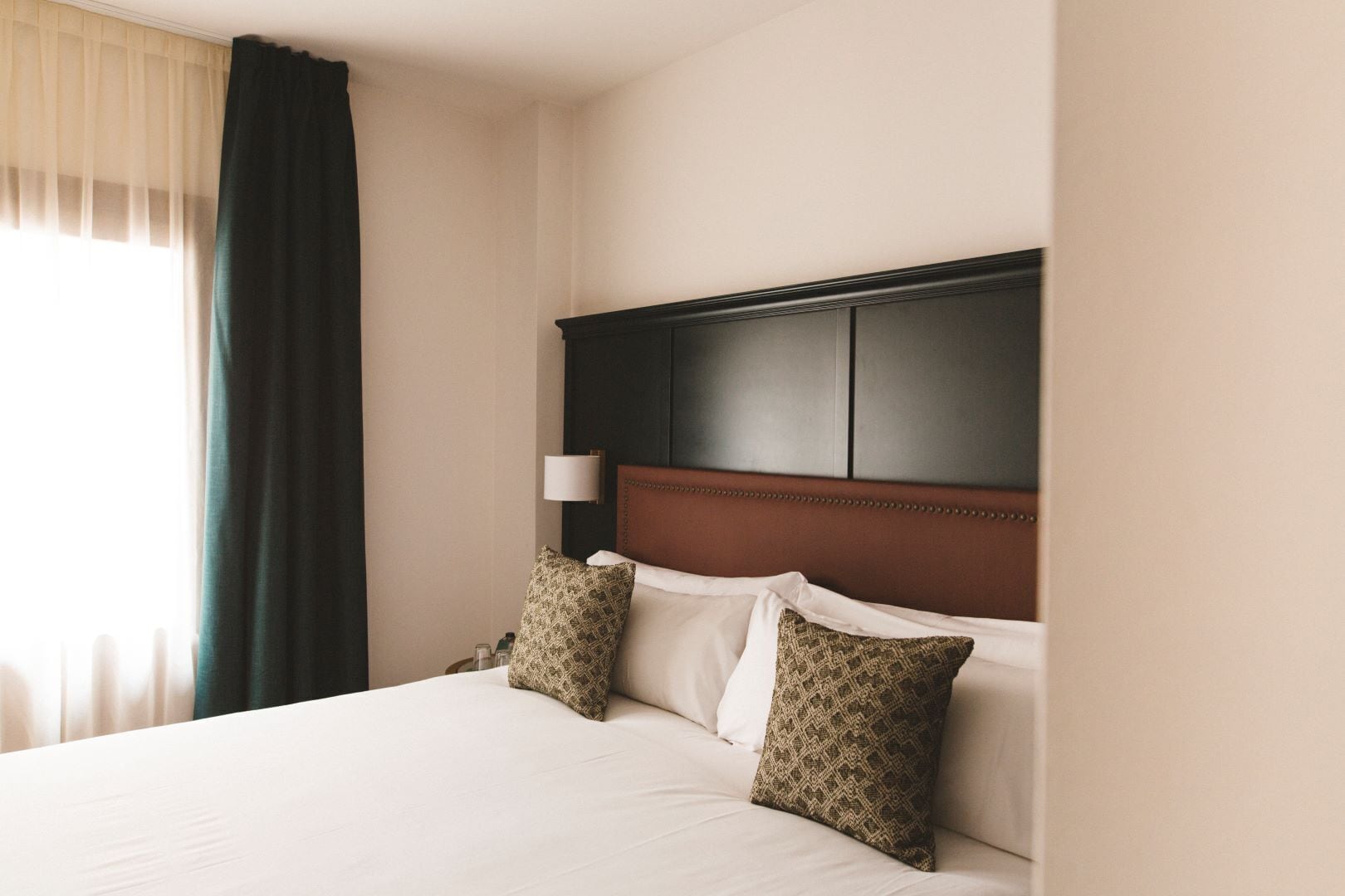 Habitación de hotel con cama, cabecero, dos almohadas, mesita de noche, lámpara y ventana con cortinas.