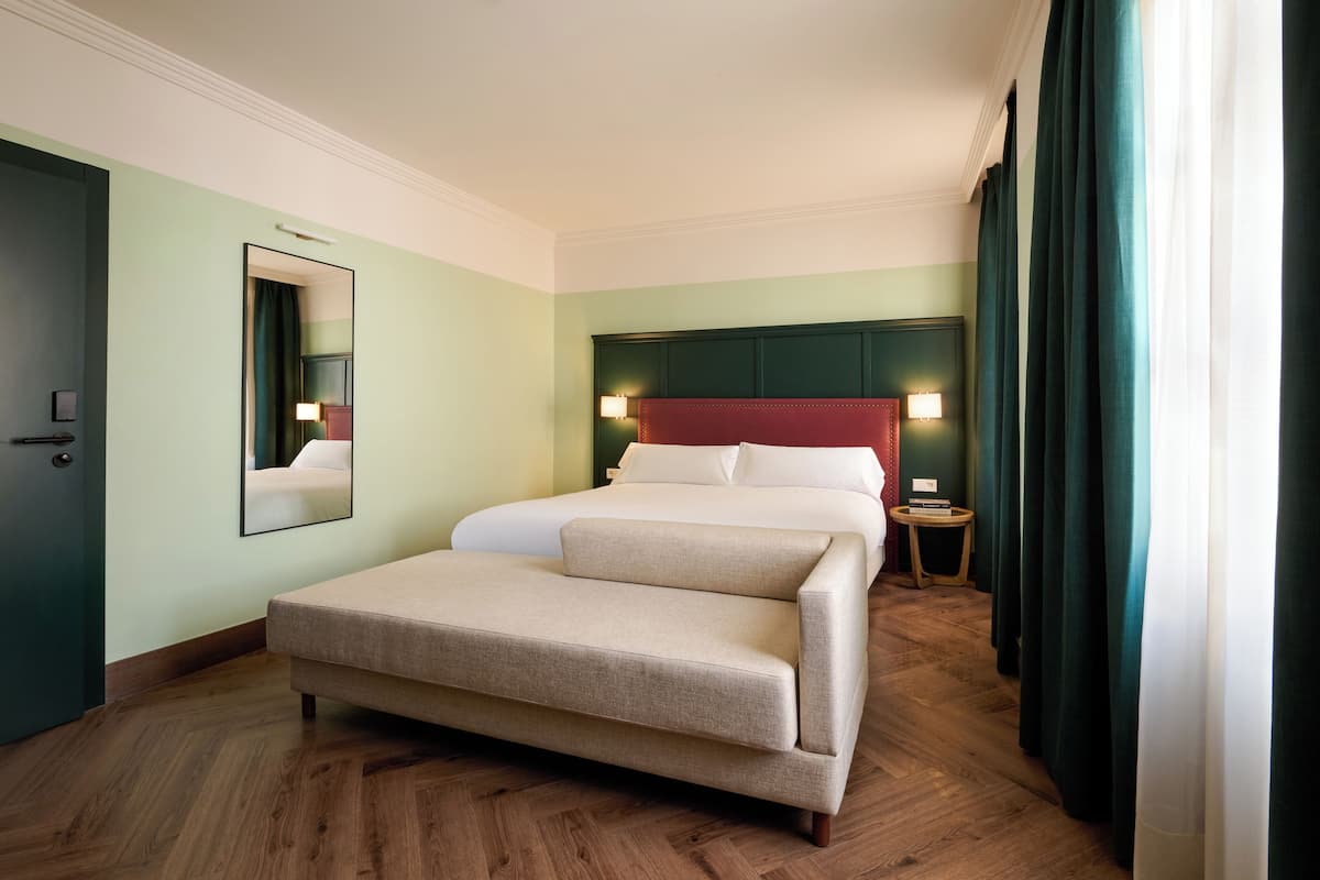 A hotel room with a bed, a sofa, a mirror, a nightstand, and a wooden floor.