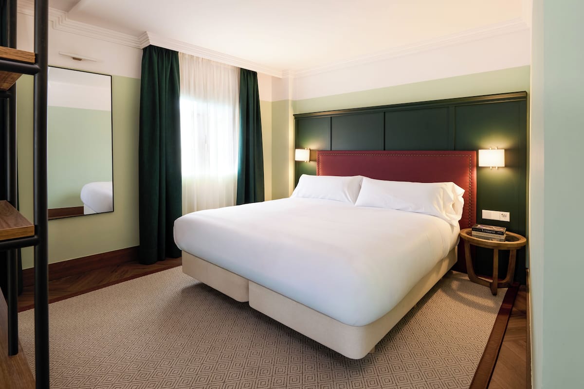 A hotel room with a large bed, a bedside table, a mirror, and a window with curtains.