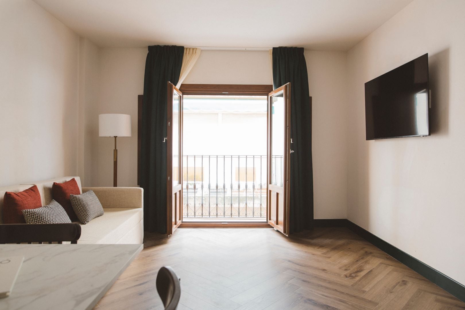 Ein Hotelzimmer mit Holzboden, einer Couch mit gemusterten Kissen, einem an der Wand montierten Fernseher und einem Balkon mit Geländer.