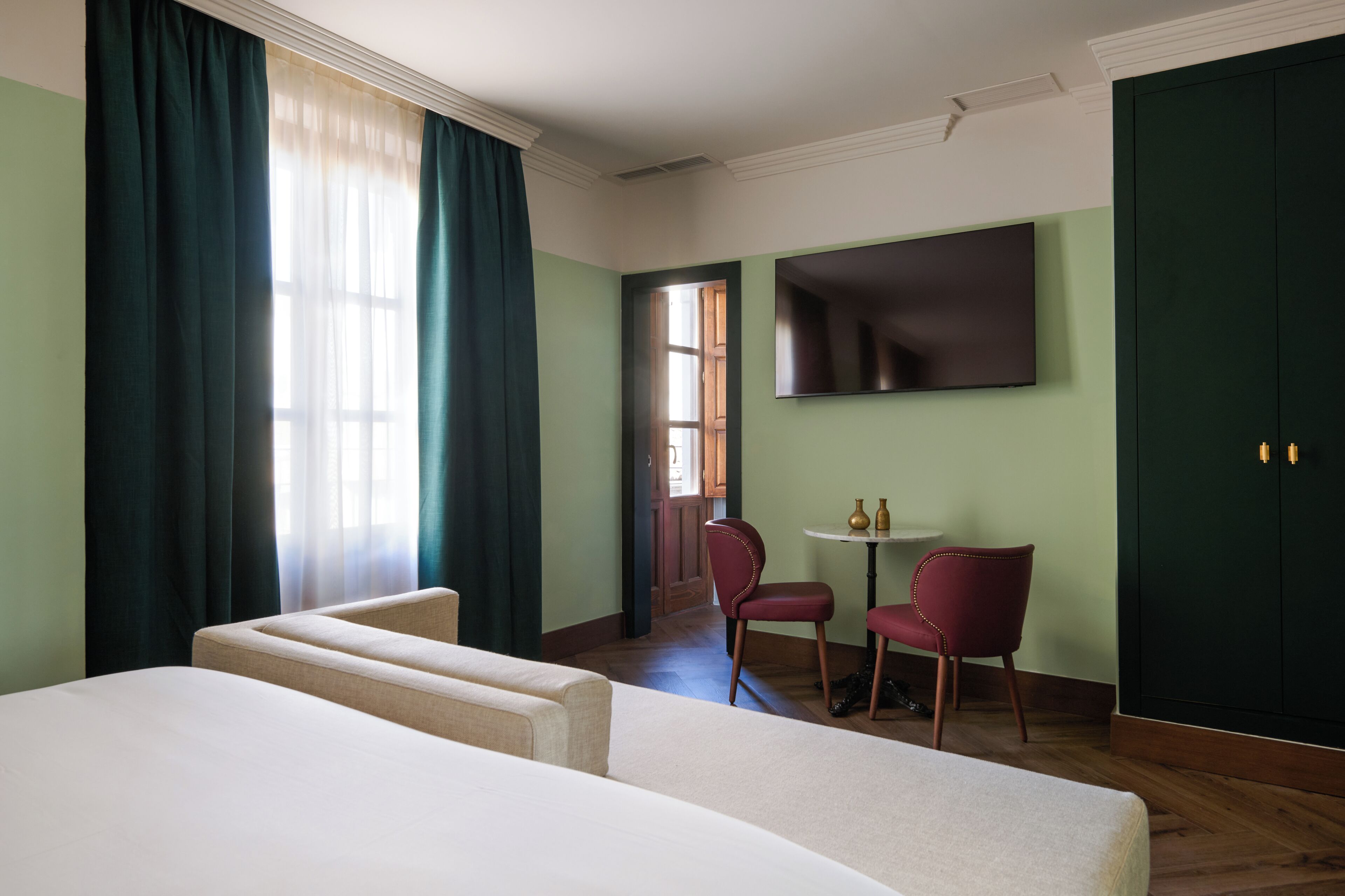 A hotel room with a bed, two chairs, a small table, and a TV on the wall.