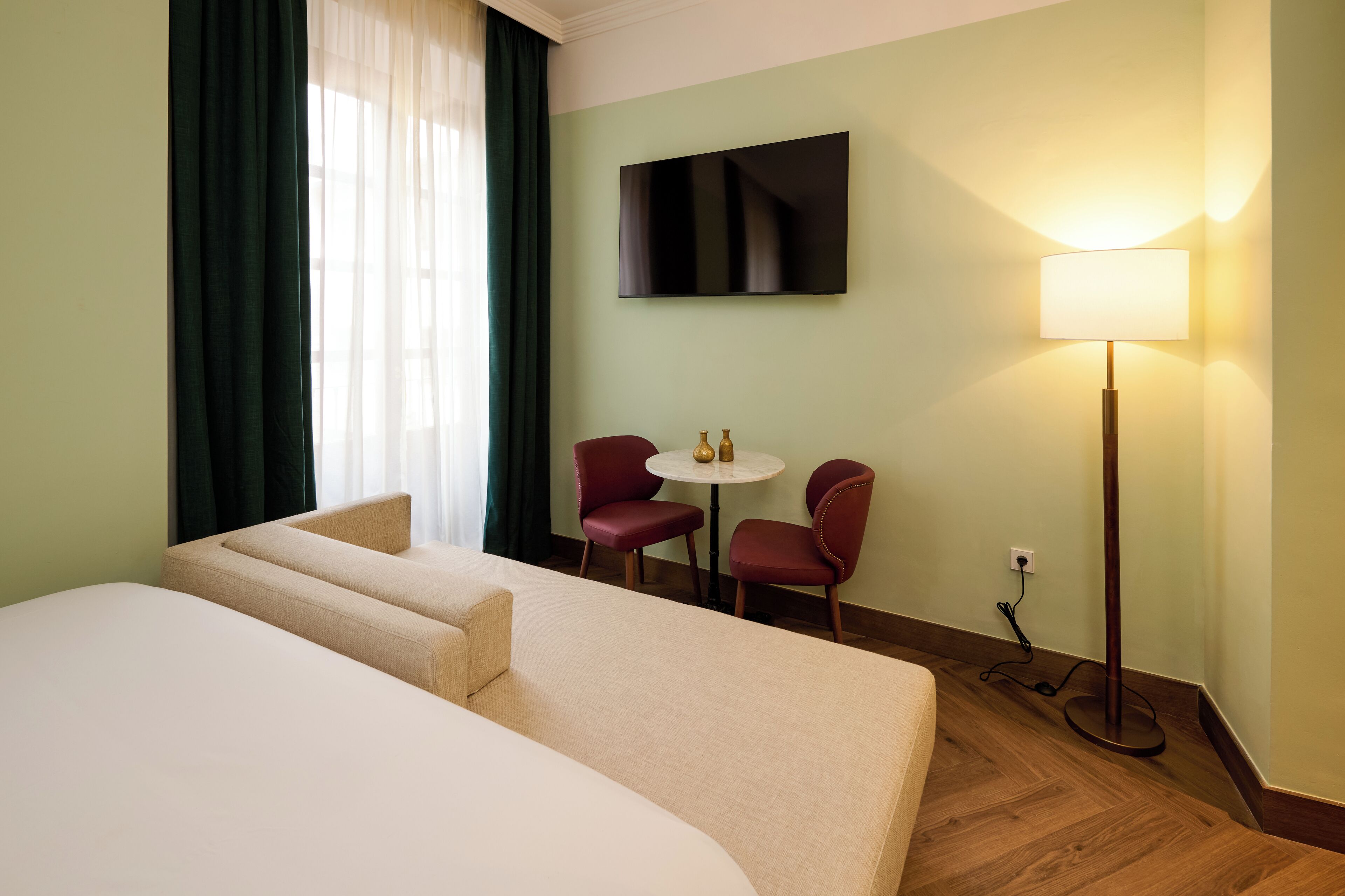 A hotel room with a large bed, a small table with two chairs, a TV mounted on the wall, and a floor lamp.