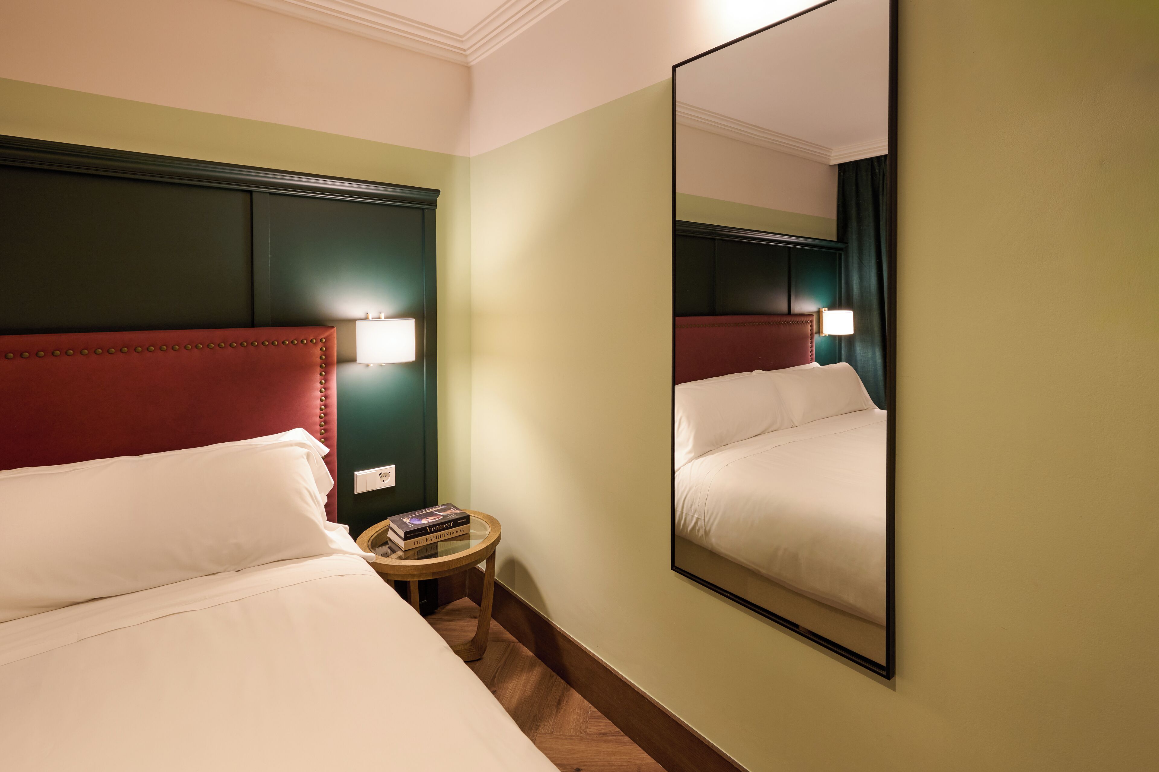 Habitación de hotel con dos camas, una mesita redonda, un espejo y una lámpara de pared.