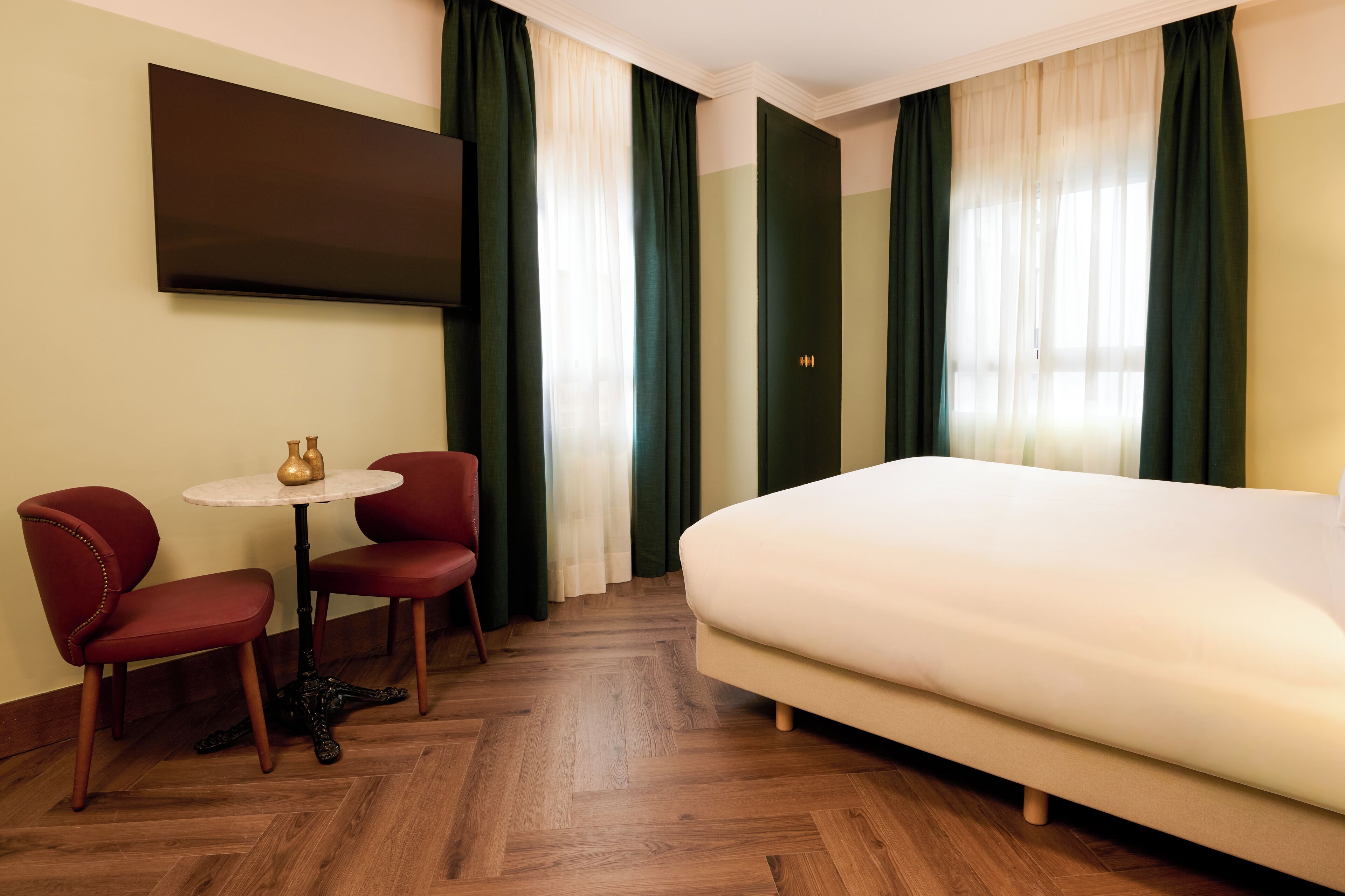 A hotel room with a large bed, two red chairs, a small round table, and a flat-screen TV mounted on the wall.