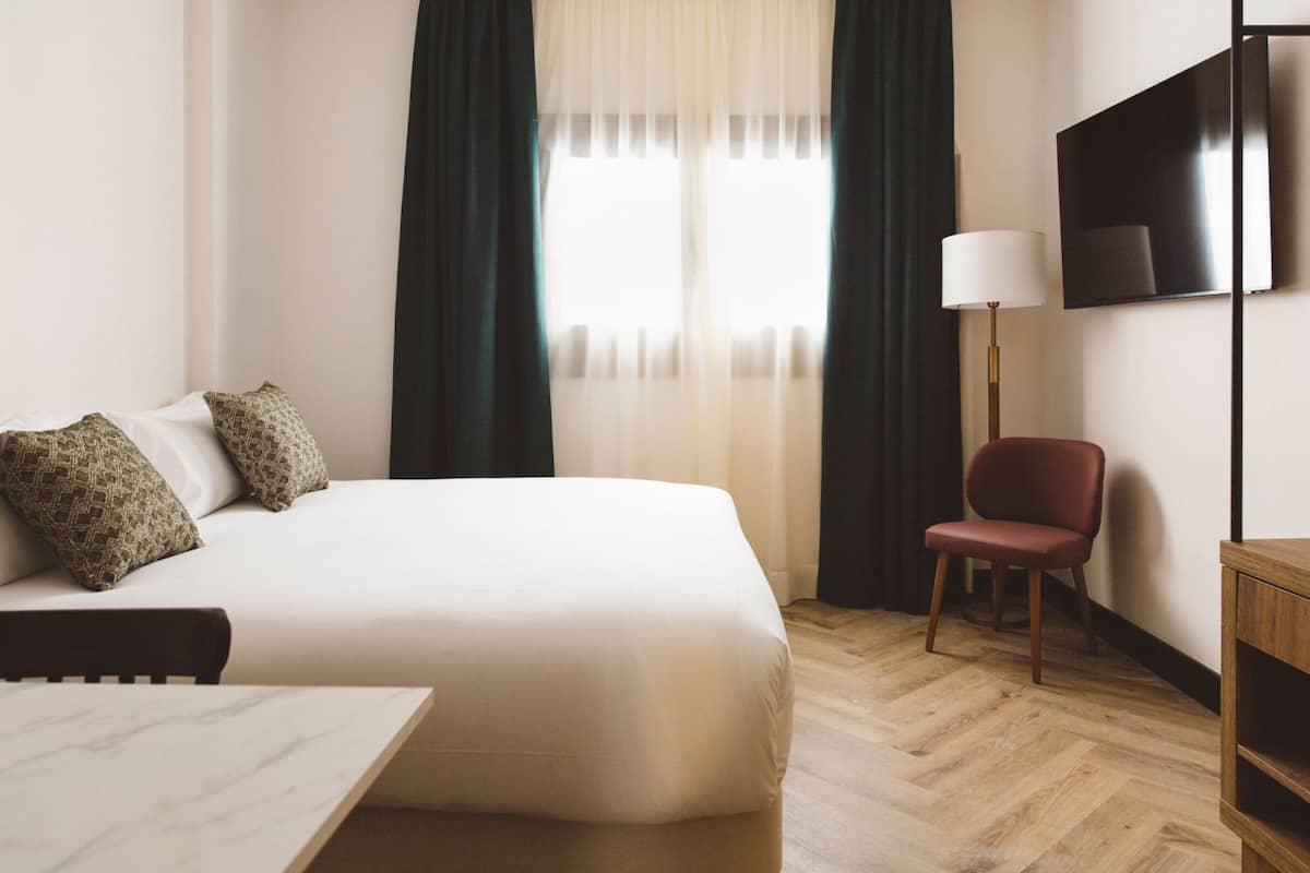 A hotel room with a bed, a chair, a desk, a TV, and a window with curtains.