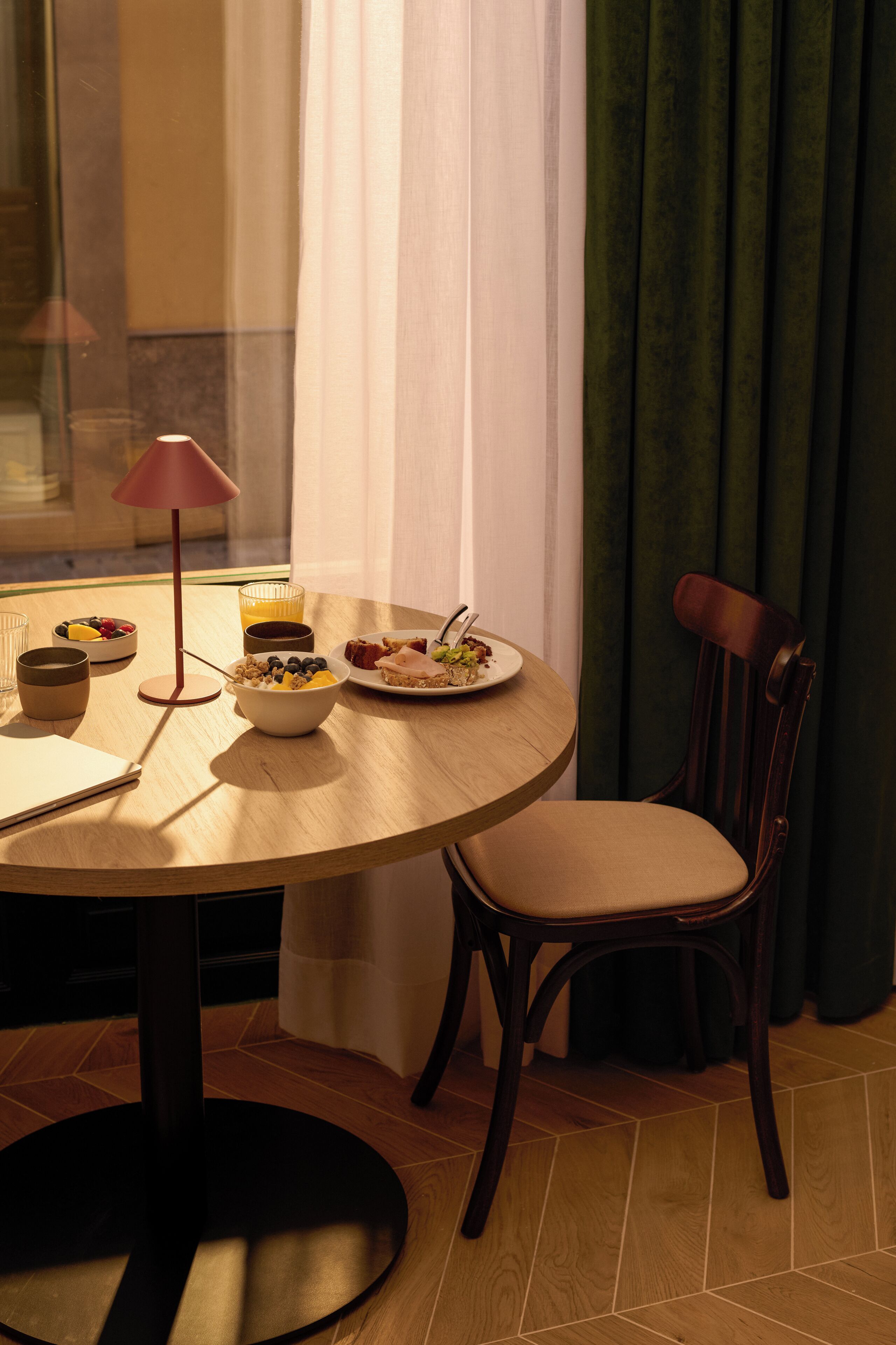 Una mesa redonda de comedor con un tazón de cereal, un plato de comida y un vaso de jugo. Hay una silla con cojín al lado de la mesa.