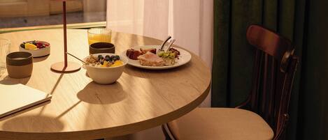 Una mesa redonda de comedor con un tazón de cereal, un plato de comida y un vaso de jugo. Hay una silla con cojín al lado de la mesa.