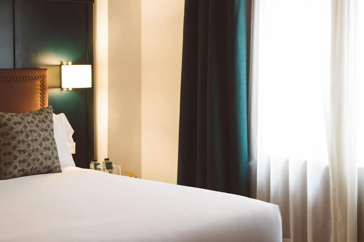 A hotel room with a bed, a headboard, a nightstand, a lamp, and a window with curtains.