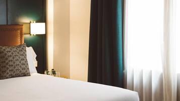 Habitación de hotel con cama, cabecero, mesita de noche, lámpara y ventana con cortinas.