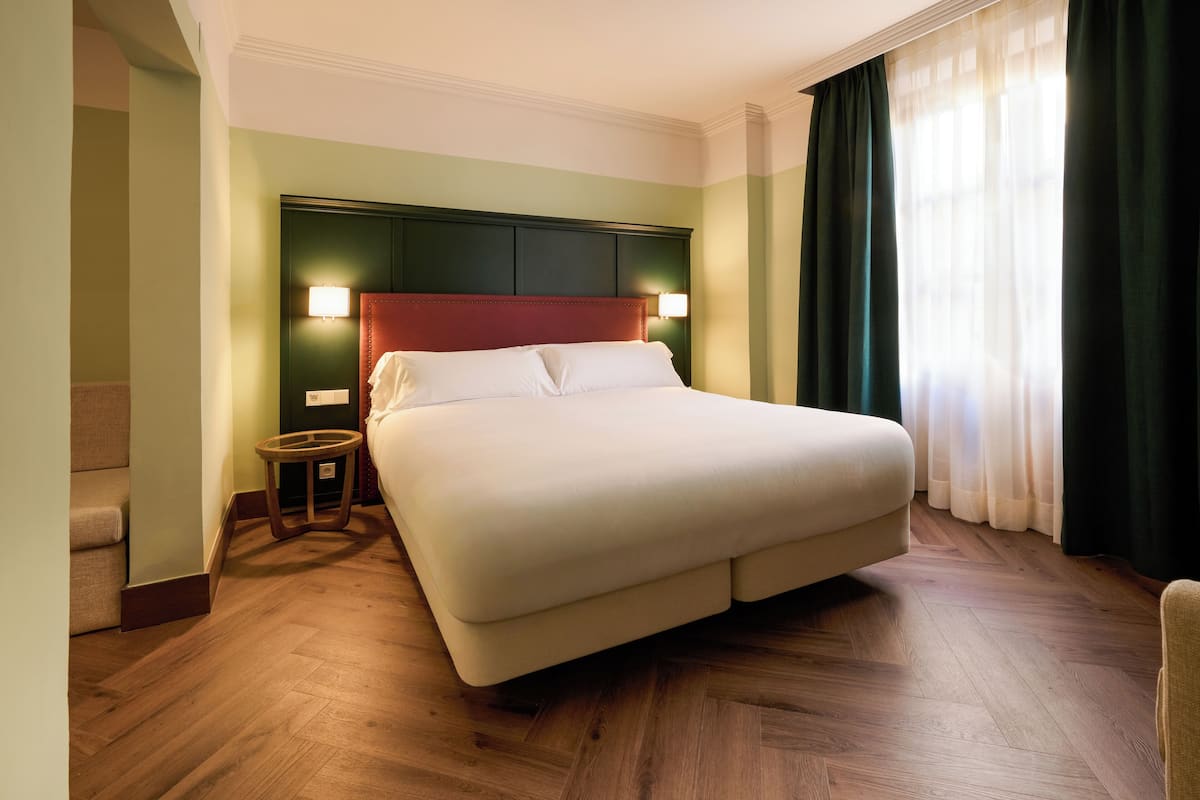 A large bed with a wooden headboard, a small round table, and a chair in a room with wooden flooring and green walls.