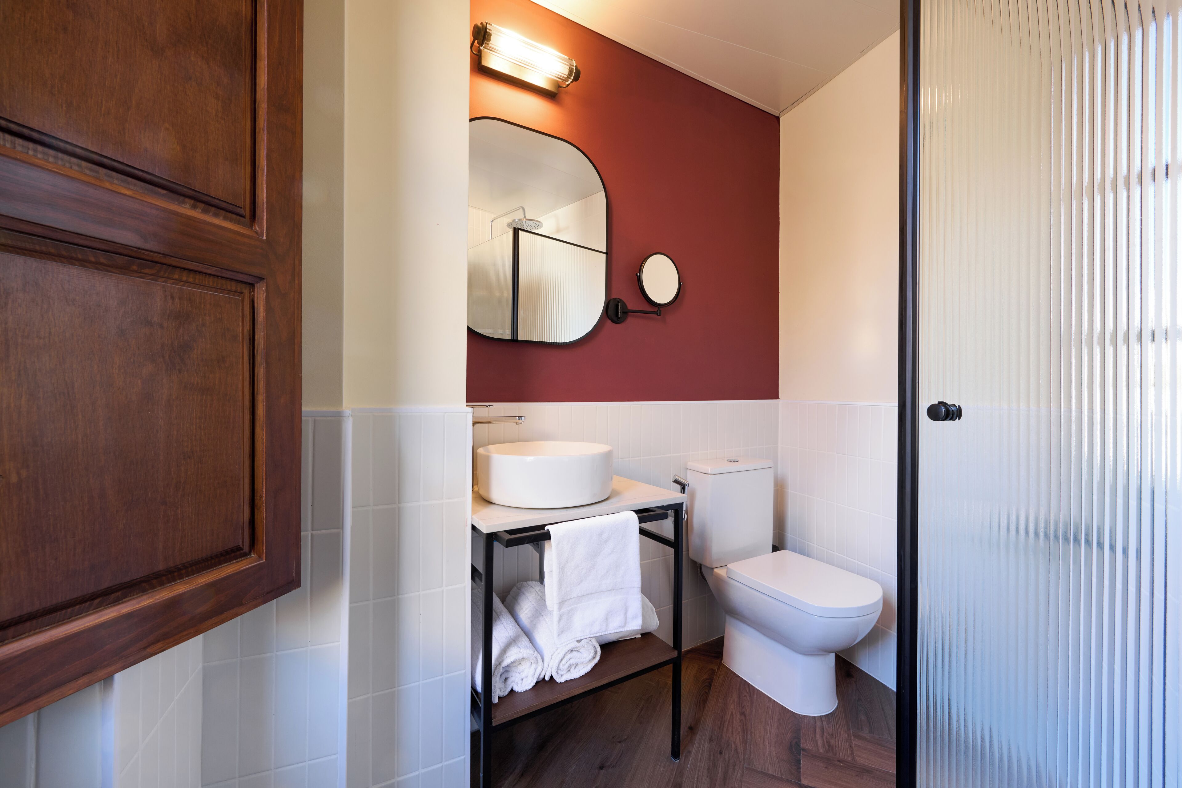 A bathroom with a wooden cabinet, a round mirror, a white sink, a toilet, and a towel rack.