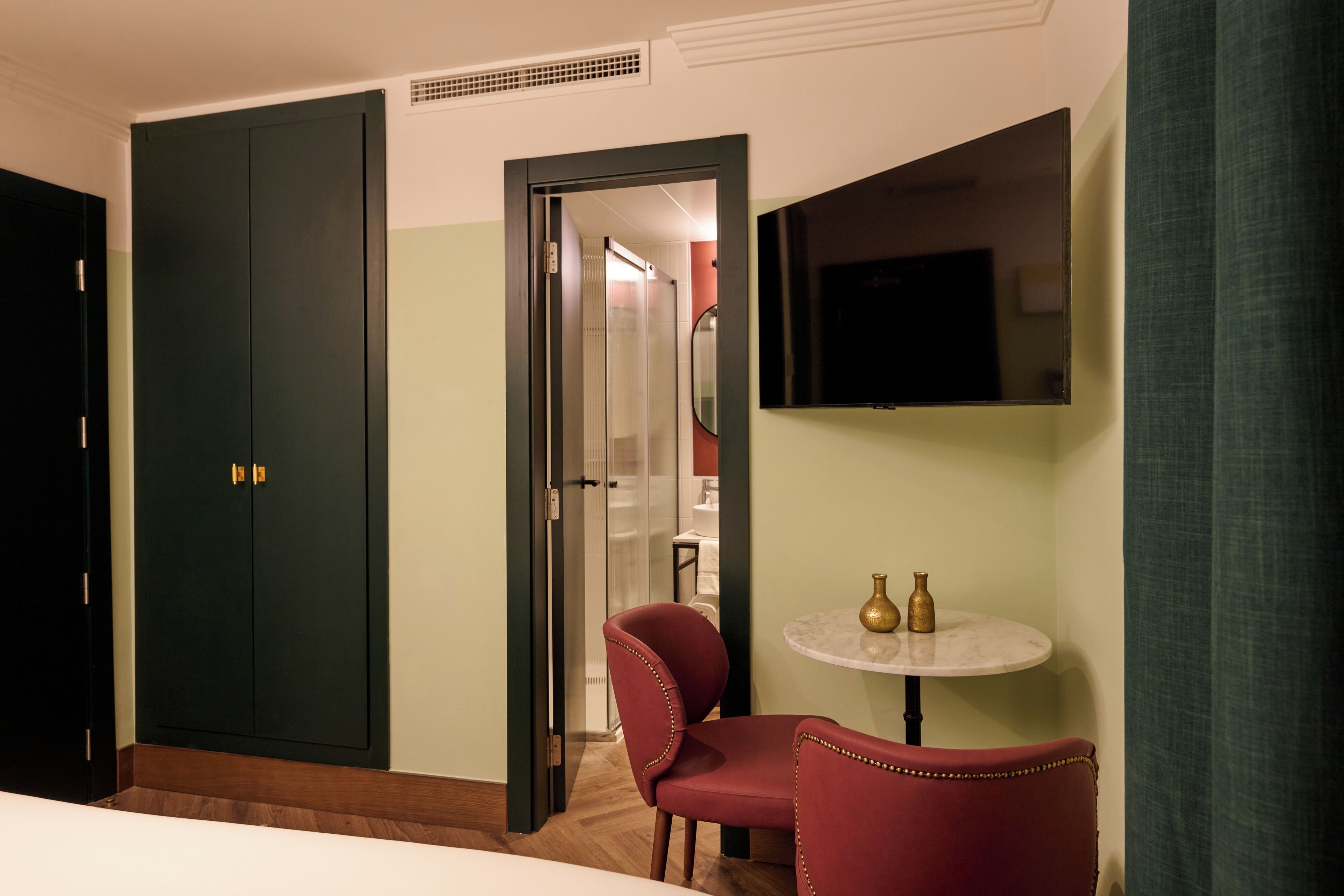 A hotel room with a bed, a red armchair, a small round table with two vases, a television mounted on the wall, and a wardrobe with a mirror.