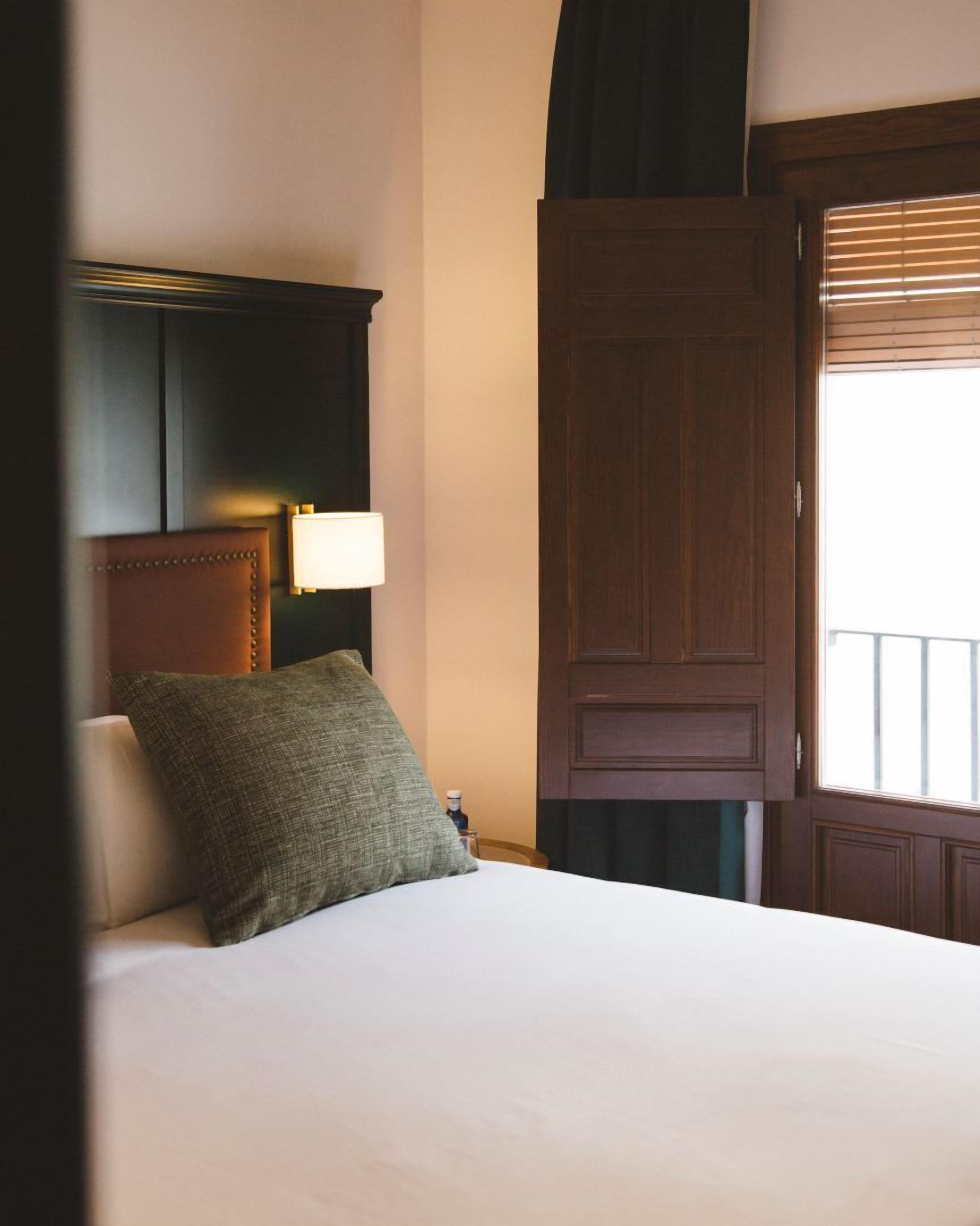 A hotel room with a bed, a nightstand, a lamp, a wooden door, and a window with blinds.