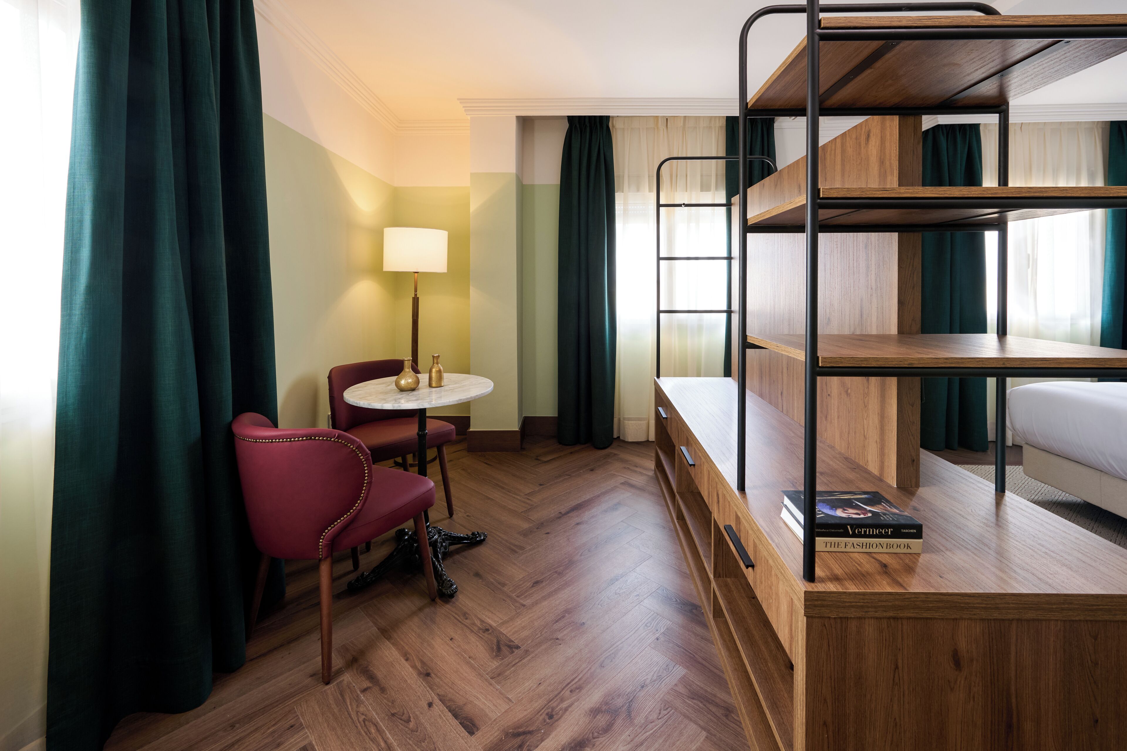 A compact hotel room with a bunk bed, a small round table with a lamp, a red chair, and a book on the bed.