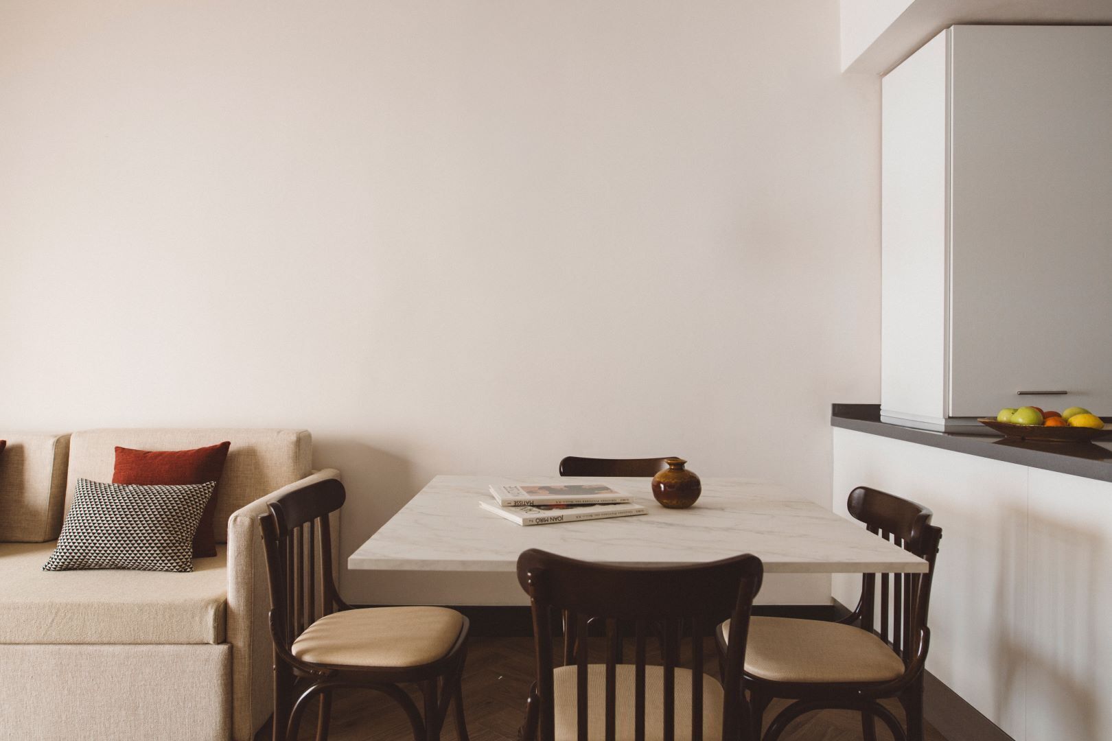 A dining area with a white table, four chairs, a vase, and a bowl of fruit on the counter.
