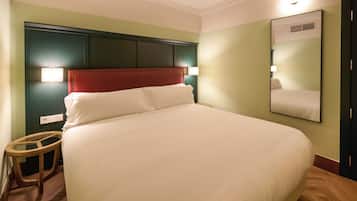 Een net opgemaakt bed met een rood hoofdeinde, een nachtkastje en een spiegel aan de muur.