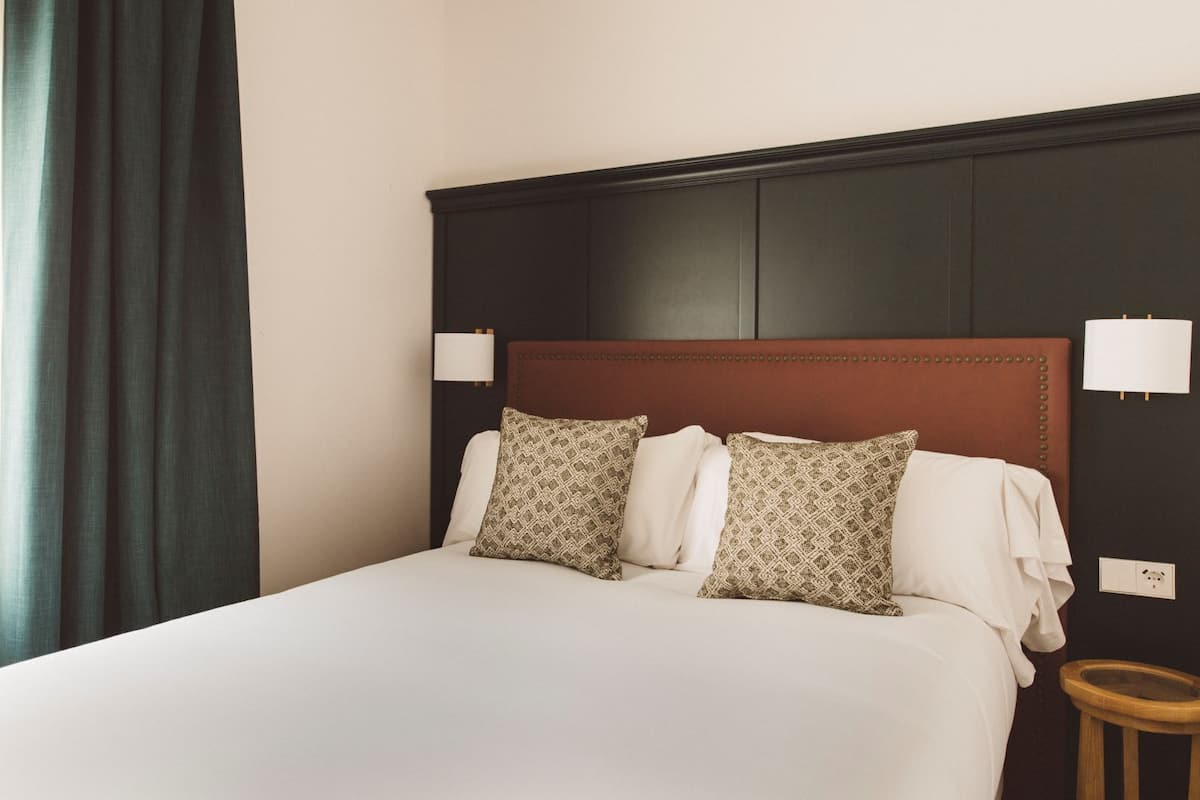 A hotel room with a large bed, two bedside lamps, a wooden nightstand, and a dark headboard.