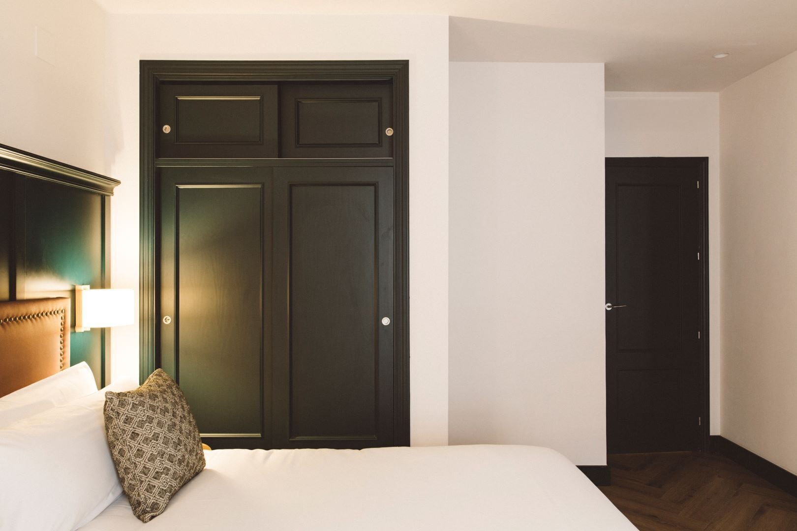 A hotel room with a large dark wooden wardrobe, a bed with white linens, and a wooden floor.