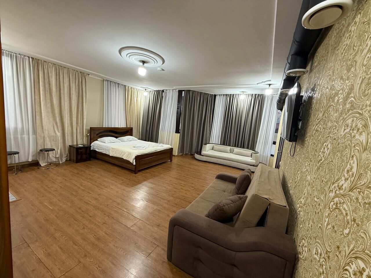 A spacious room with a wooden floor, a bed, a sofa, and a TV mounted on the wall.