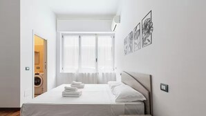 Minimalistinen hotellihuone, jossa on sänky, puinen yöpöytä, ikkuna, jossa on läpikuultavat verhot, ja seinälle asennettu ilmastointilaite.