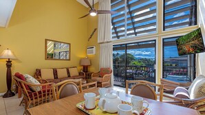 A room with a dining table set for tea, wicker furniture, a ceiling fan, and a large window with a view of the outdoors. - Ocean view loft with pool and hot tub near Kihei beaches summer deals (Kihei)