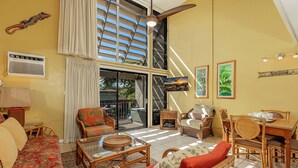 A spacious living area with a high ceiling, large windows, and a view of the outdoors. There is a dining area with wooden furniture and a television mounted on the wall. - Ocean view loft with pool and hot tub near Kihei beaches summer deals (Kihei)