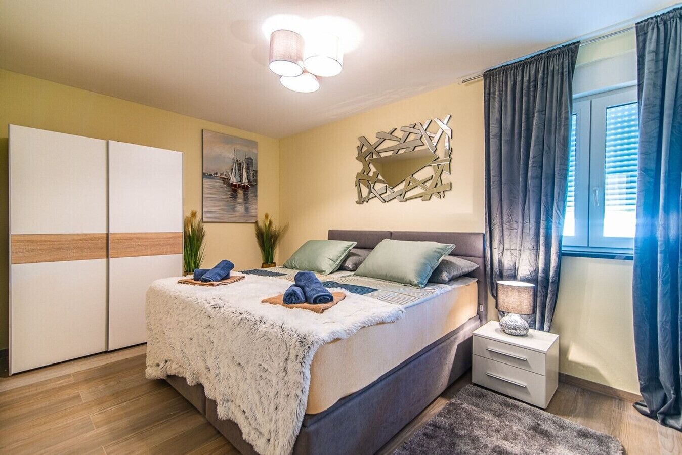 Une chambre à coucher comprenant un lit, des tables de chevet, une commode, une fenêtre avec des rideaux et une œuvre d’art accrochée au mur.