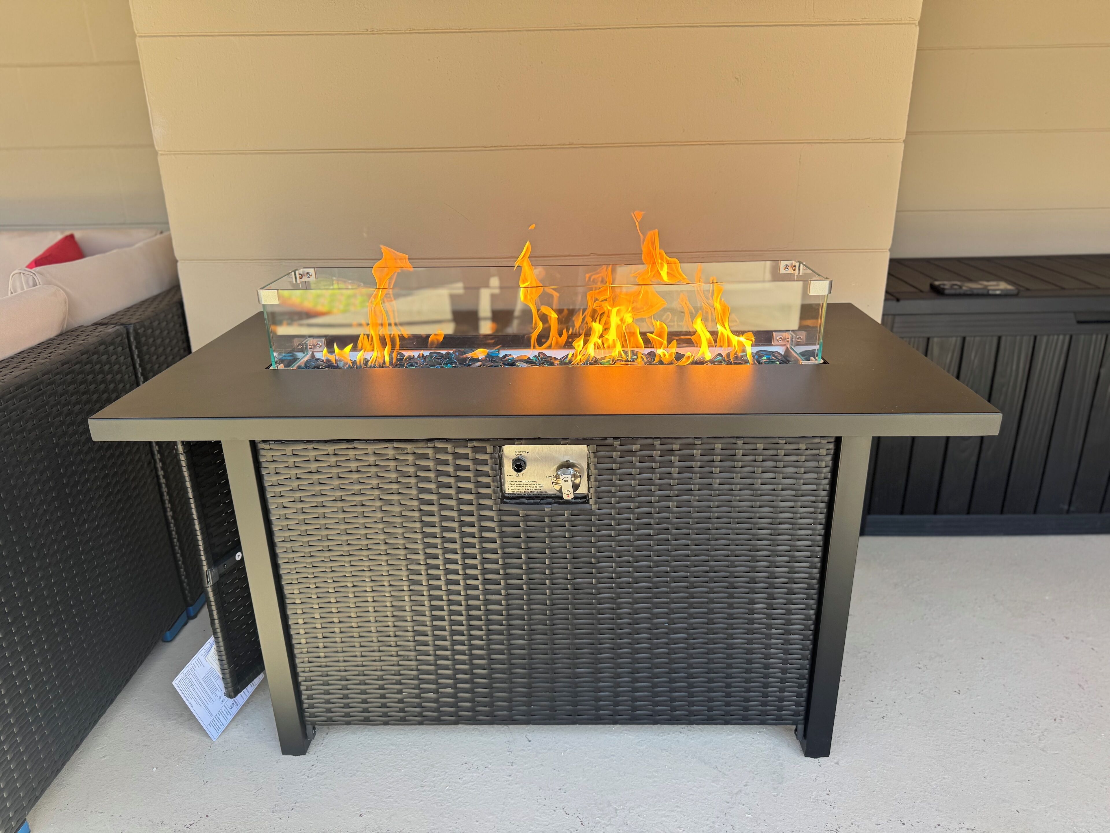 A rectangular outdoor fire feature with a metal frame and glass top, surrounded by a wicker base.