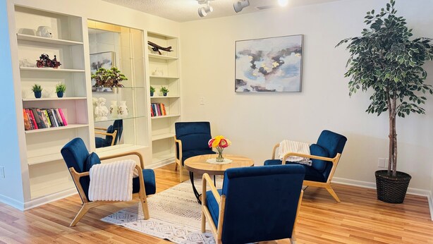 A living room with a wooden floor, blue armchairs, a round table with a vase of flowers, a bookshelf, and a potted plant. - 5/3 Pool Home In Central Lakeland, Pet Friendly! (Lakeland)