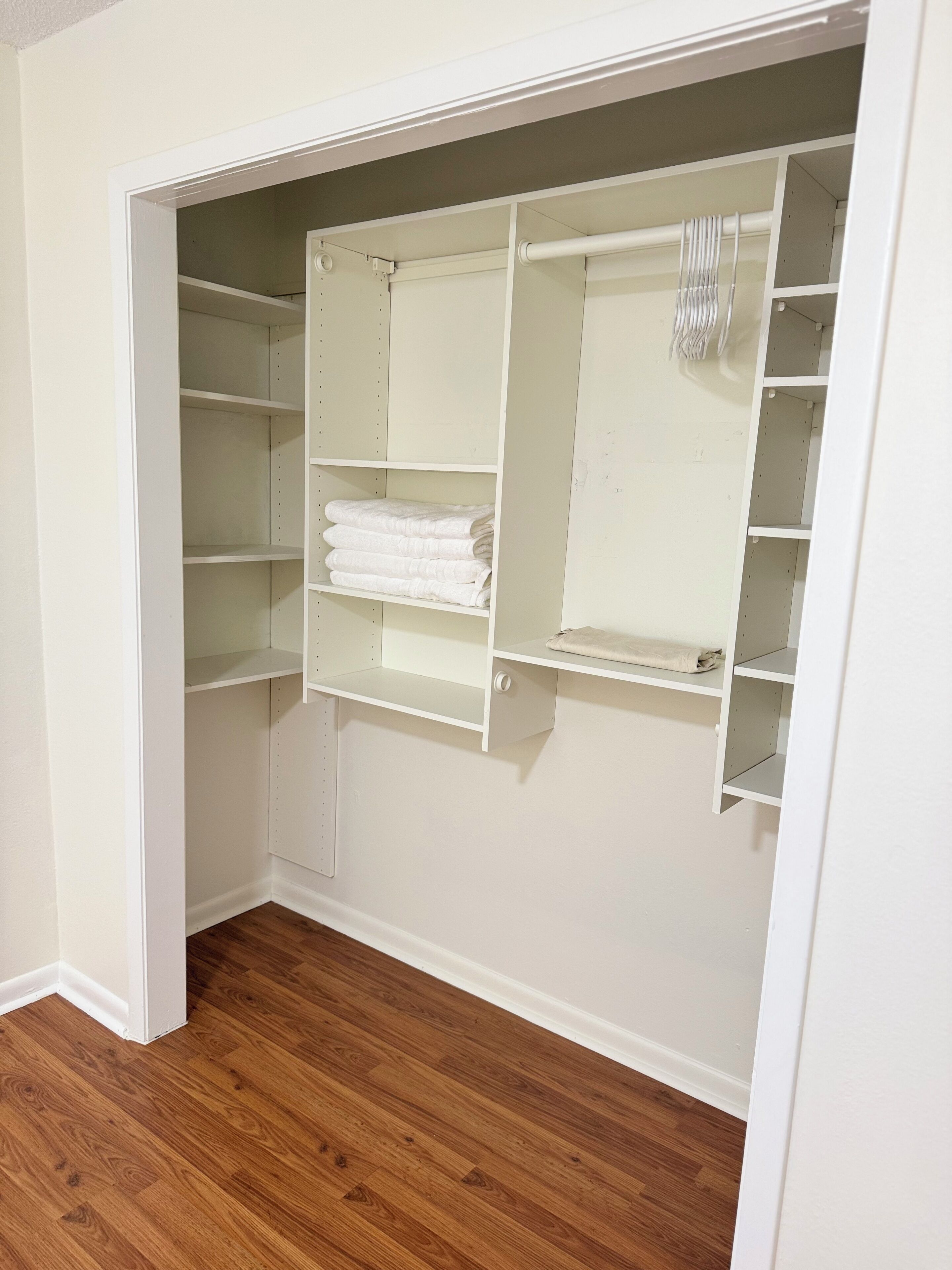 A walk-in closet with multiple shelves and hanging space, wooden flooring, and white walls.