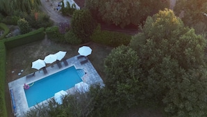 Vista aérea de uma propriedade com piscina, espreguiçadeiras e guarda-sóis. Vários edifícios exibem um estilo arquitetônico tradicional.