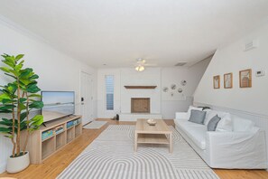 A modern living room with a white couch, a wooden coffee table, a TV on a wooden stand, a fireplace, and a large plant. - 3-bedroom house in marvelous North Myrtle Beach - Cozy and Modern (North Myrtle Beach)