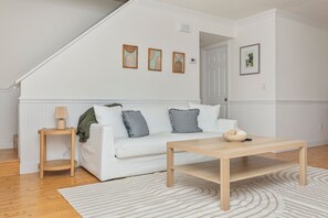 A white living room with a wooden coffee table, a white sofa with grey cushions, a small wooden side table with a lamp, and a white rug. - 3-bedroom house in marvelous North Myrtle Beach - Cozy and Modern (North Myrtle Beach)