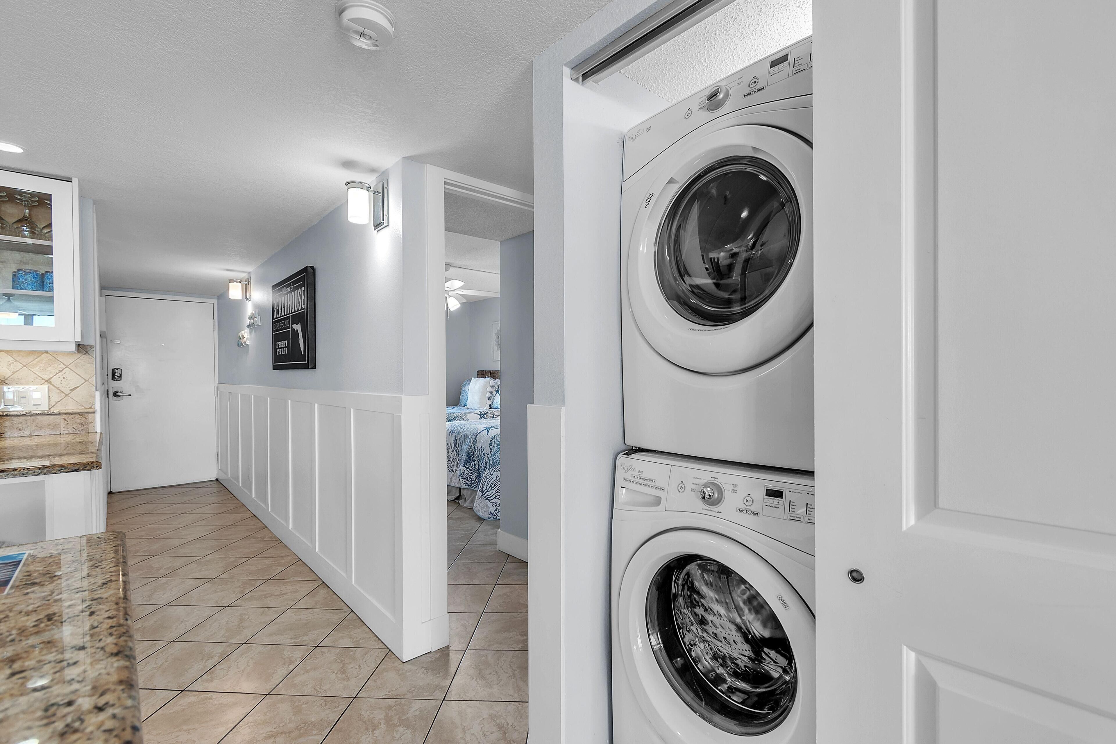 A laundry room with two stacked front-loading washers and dryers, a tiled floor, and a hallway leading to a bedroom.
