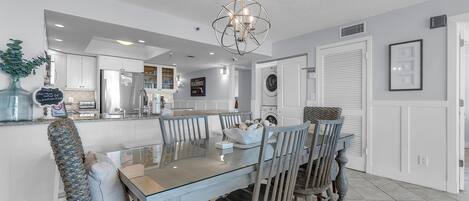 Una cocina moderna con armarios blancos, un refrigerador de acero inoxidable y una zona de comedor con mesa de vidrio y sillas de madera.