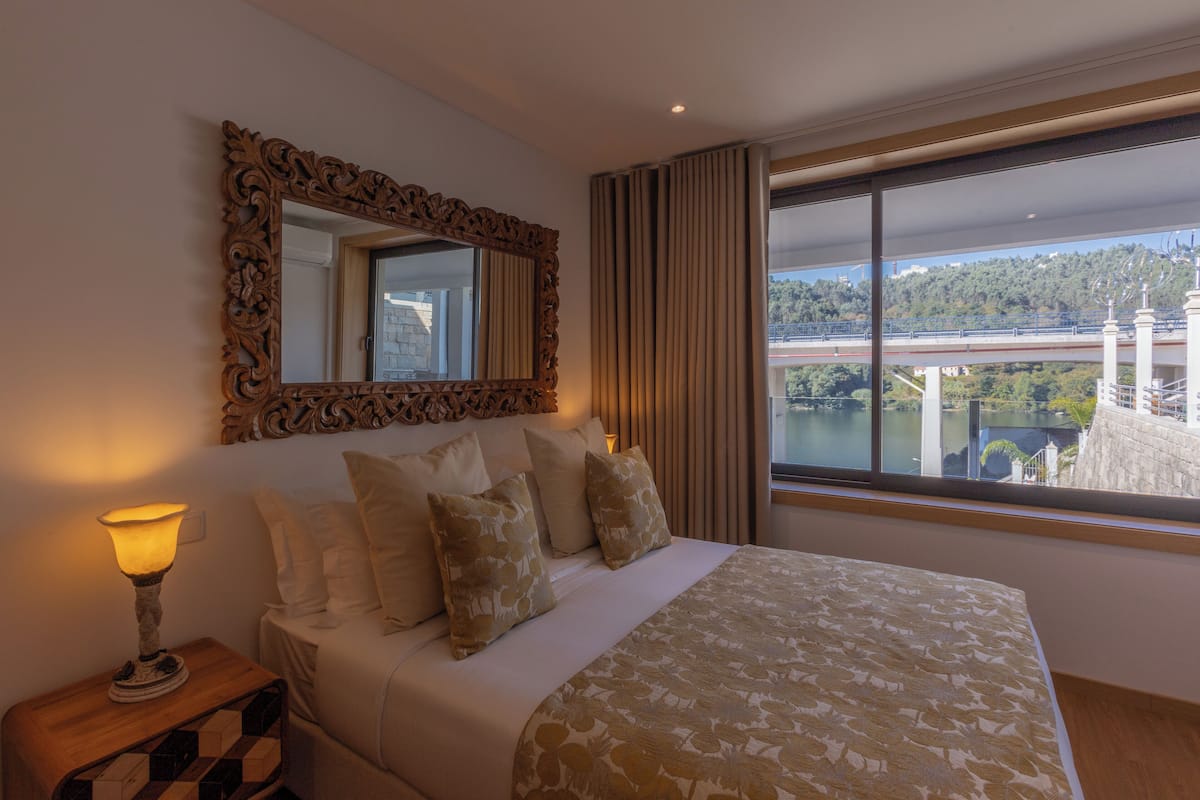 A hotel room with a large bed, a mirror, a nightstand with a lamp, and a window with a view of a bridge and greenery.