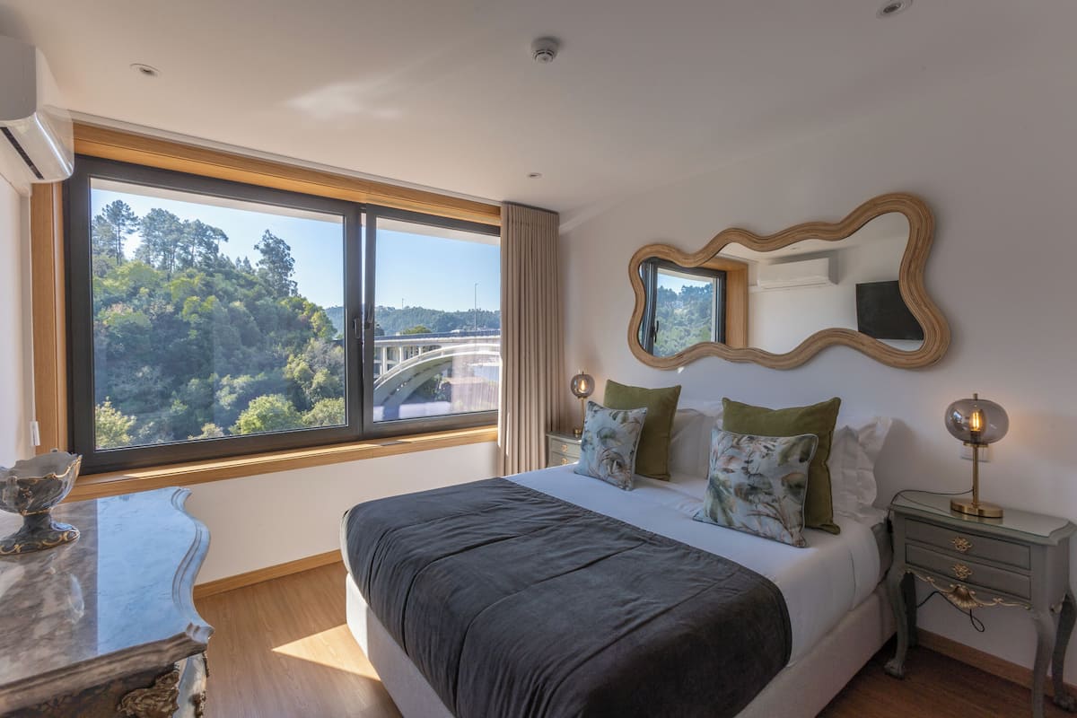 A bedroom with a large window, a bed with multiple pillows, a nightstand with a lamp, and a marble table.