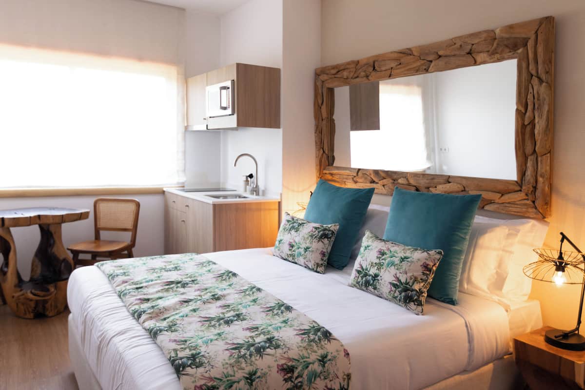 A bedroom with a bed, pillows, a bedside lamp, a wooden headboard, a large mirror, a window, and a kitchenette with a sink and cabinets.