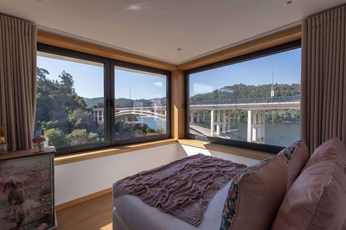 A bedroom with a large window offering a view of a bridge and greenery.