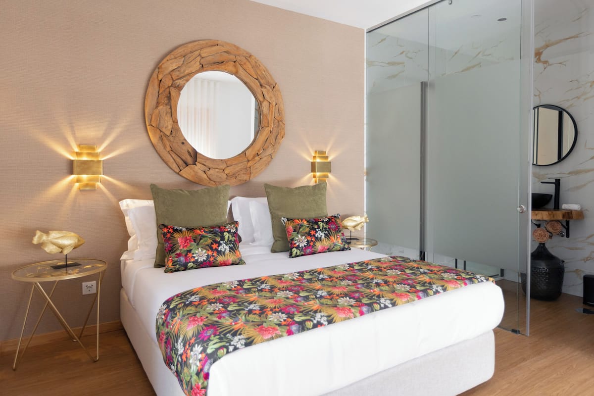 A modern bedroom with a large bed, two wall-mounted lights, a round mirror, and a glass shower door.