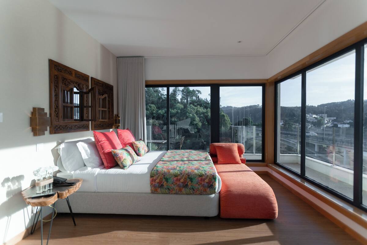 A modern bedroom with a large bed, a small table, and a view of the city through large windows.
